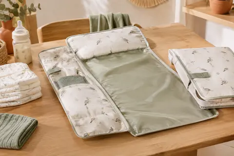 Matelas à langer nomade pliable avec coussin et poches, posé près de couches pliées sur une table en bois.